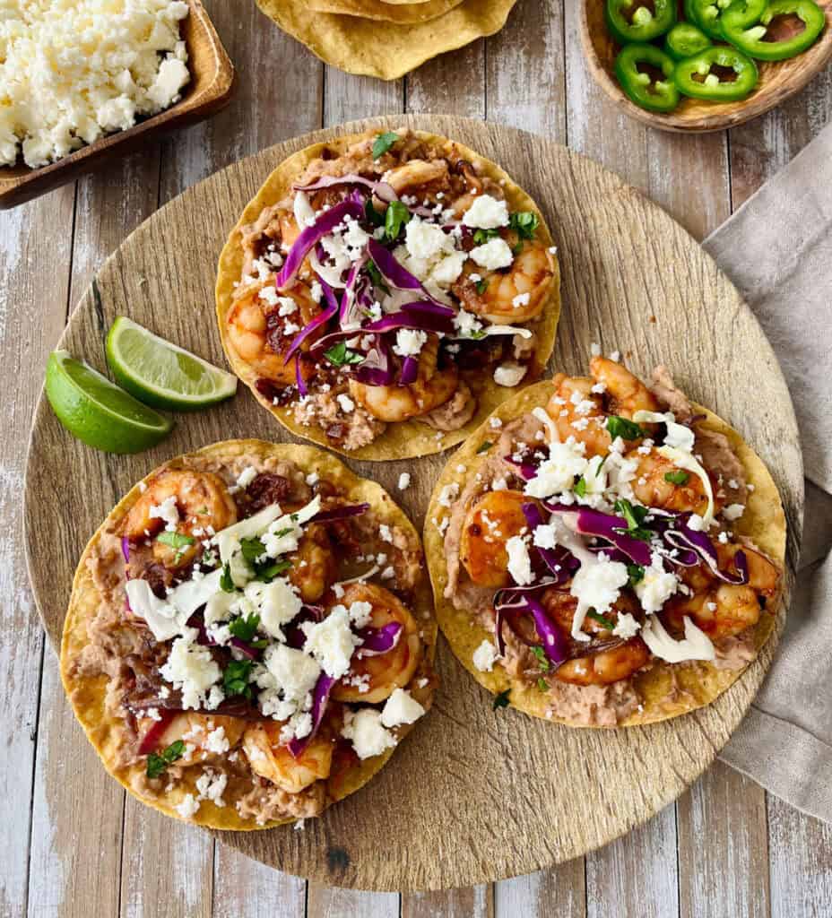 Three shrimp tostadas topped with cheese, onions, and sliced limes on a wooden serving board with dishes of crumbled cheese, tostadas, and sliced jalapeño peppers.