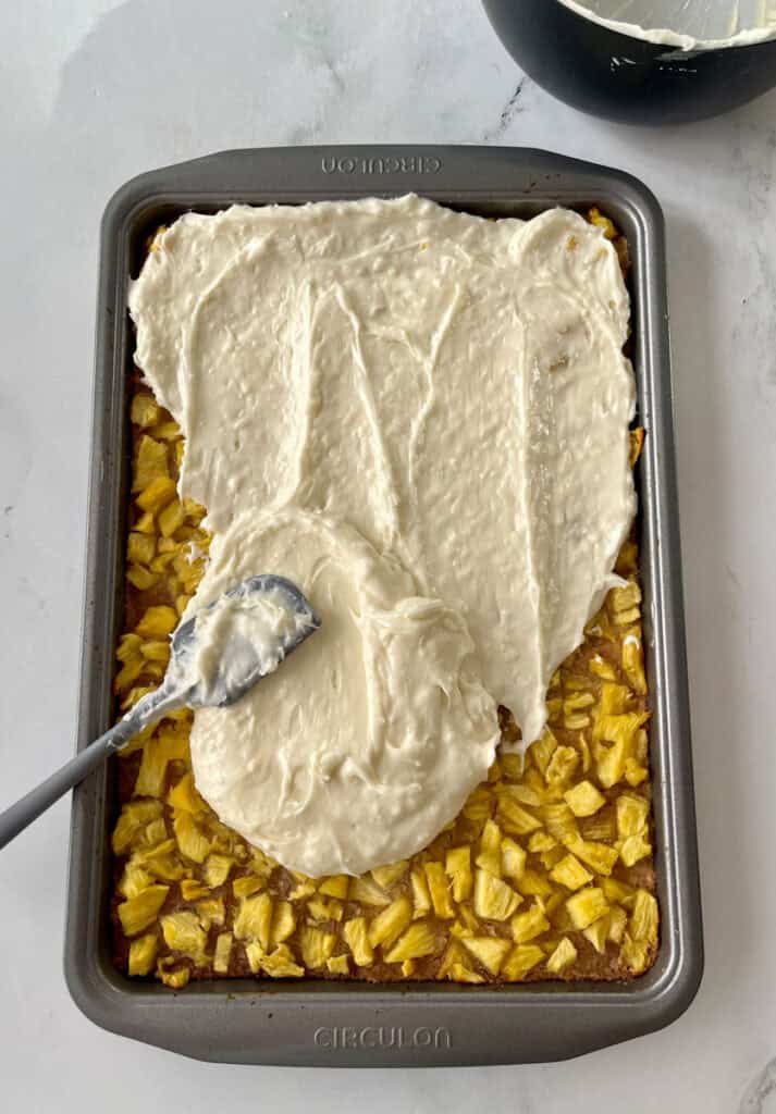 A baking pan filled with a baked crust and a pineapple filling topped with a layer of coconut frosting on a white surface.