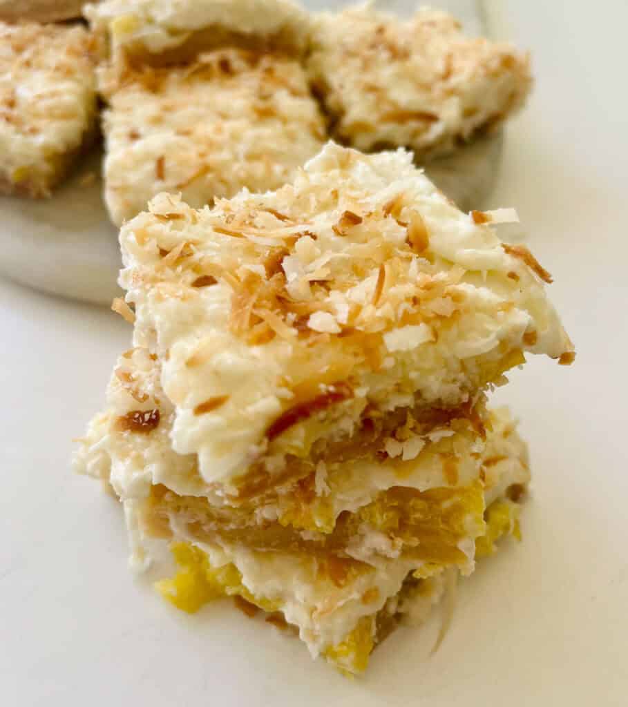 Delicious homemade pineapple bars with coconut cream frosting bars with toasted coconut topping sitting on a white surface.
