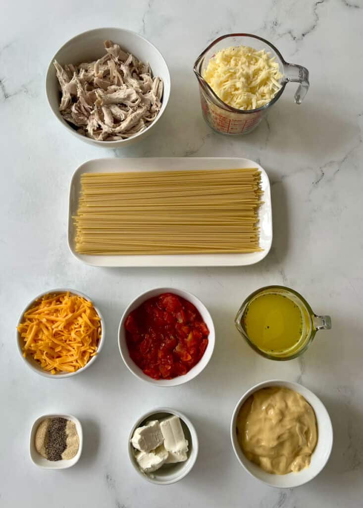 Uncooked spaghetti with shredded chicken, cheese, diced tomatoes, condensed soup, broth, and seasonings on a marble surface.