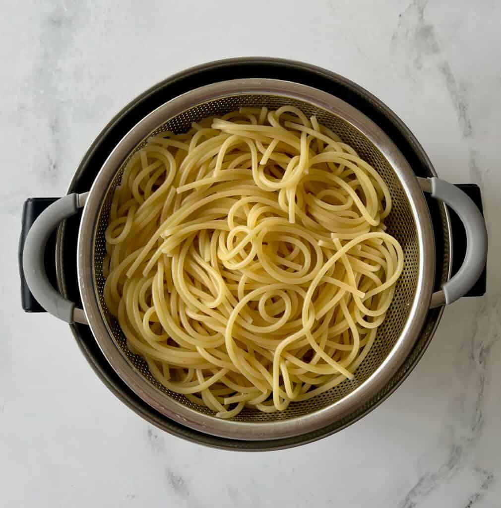 Cooked spaghetti pasta in a metal strainer on a white surface.