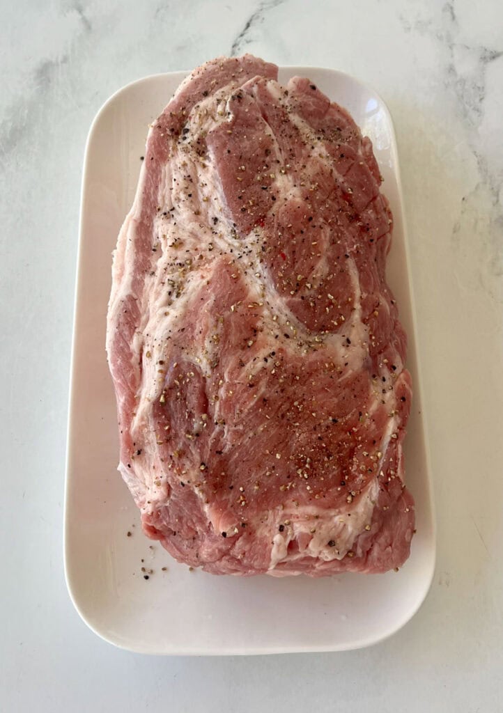 Raw pork roast seasoned with salt and pepper on a white plate.