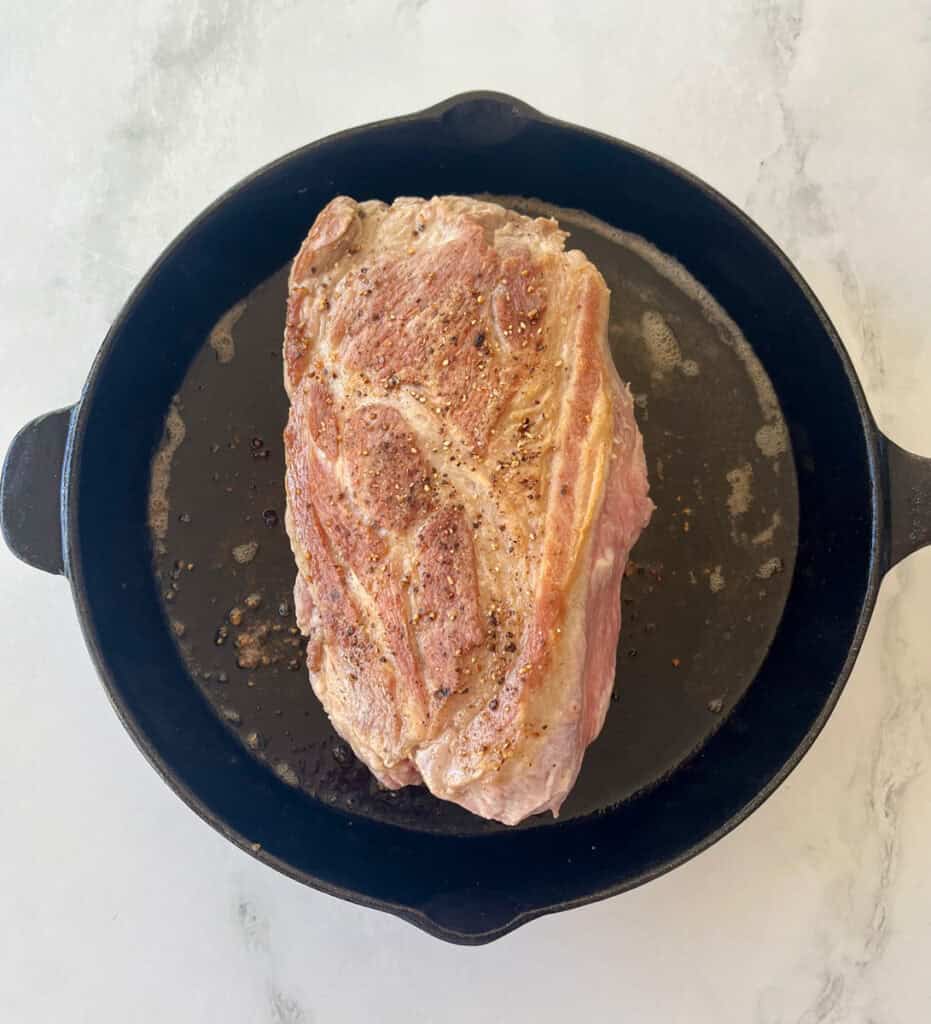 Raw pork roast seasoned and cooking in a black skillet on a white surface.