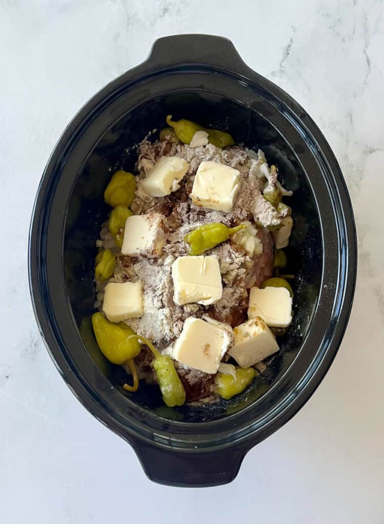 Slow cooker pork roast with pepperoncini peppers, chunks of butter, and seasonings in a black crockpot.
