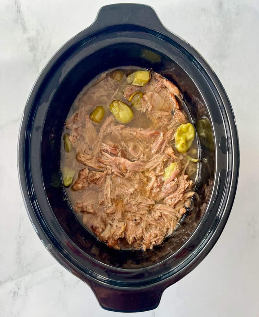 Tender pulled pork in slow cooker with pepperoncini peppers and juices.