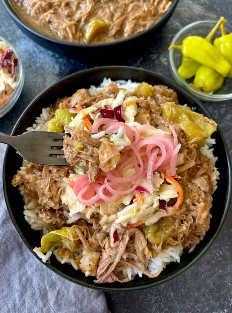 Slow cooker pulled pork served over rice with coleslaw and pickled onions. Bowls of peppers and coleslaw on the side.