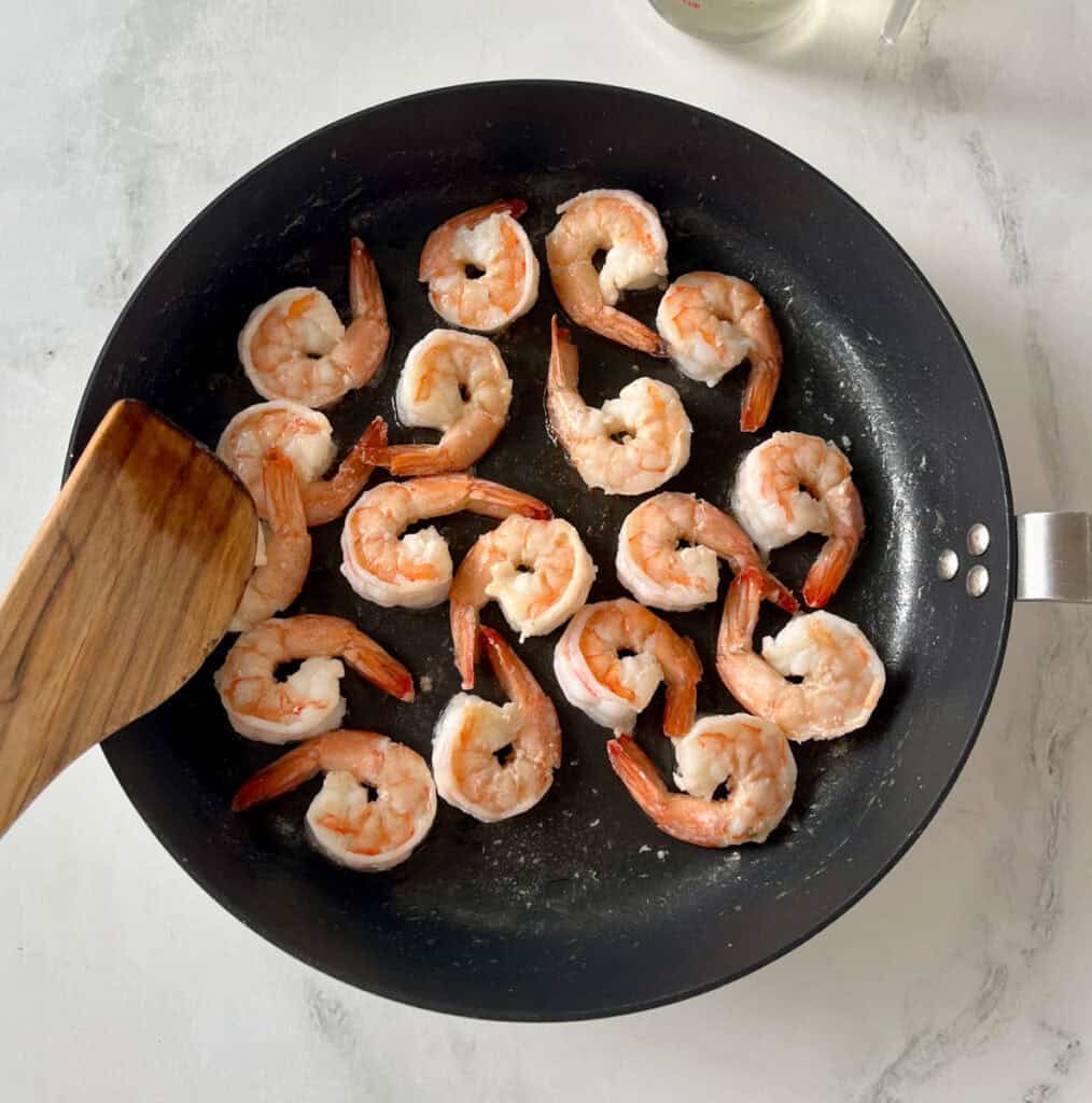 Shrimp cooking in a black skillet with a wooden spatula.