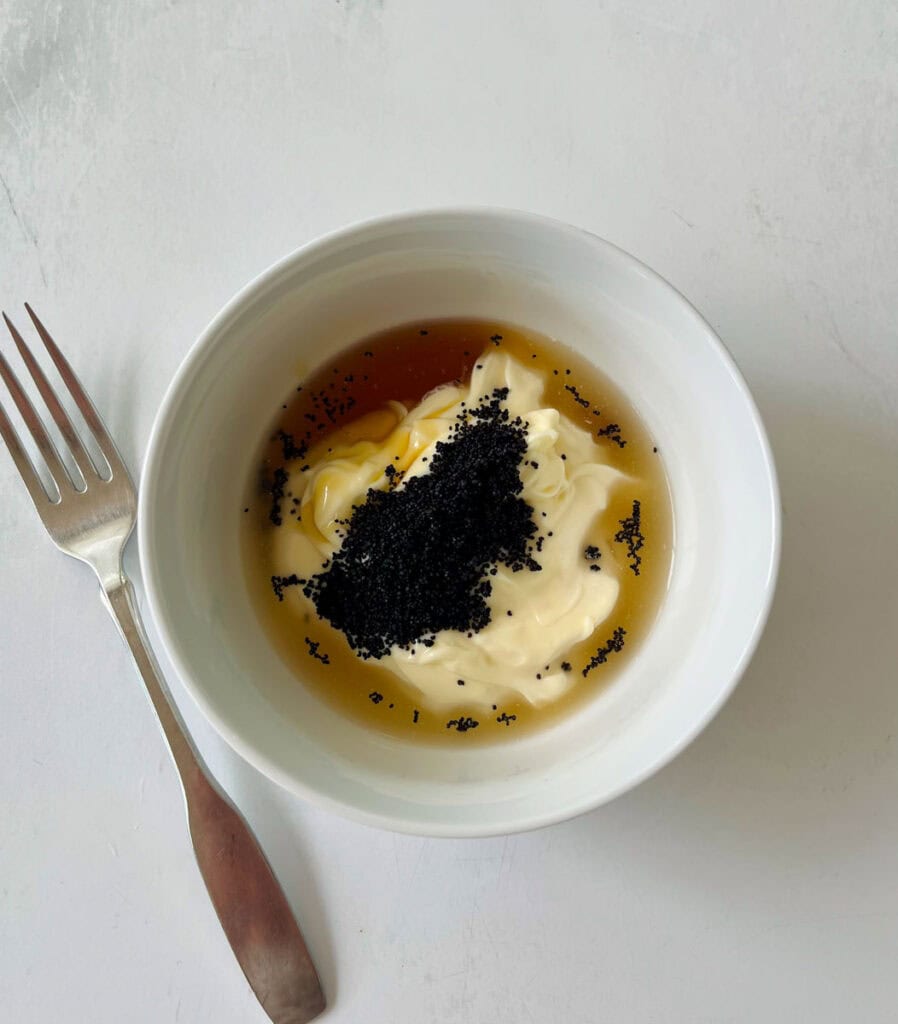 A white bowl filled with mayonnaise topped with black poppy seeds, cider vinegar, and honey, placed on a white surface with a fork beside it.