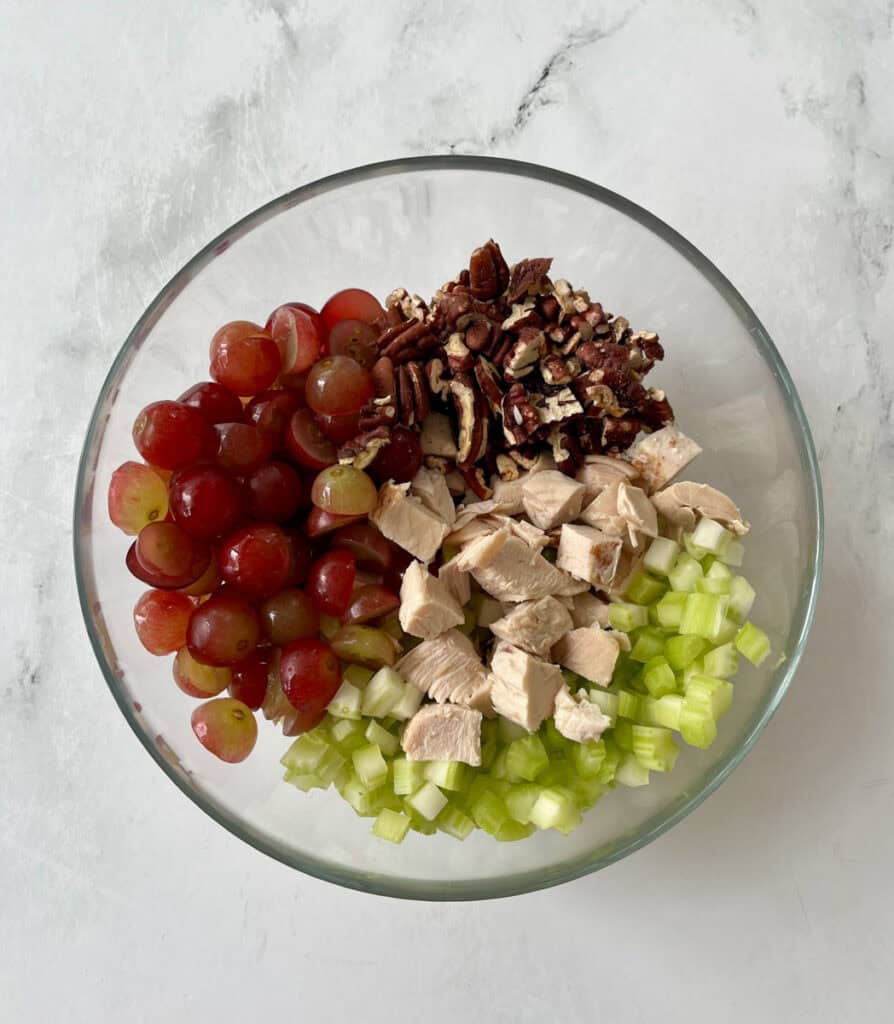 A glass bowl containing fresh grapes, cooked chicken pieces, chopped pecans, and diced celery, ready for making a chicken salad.