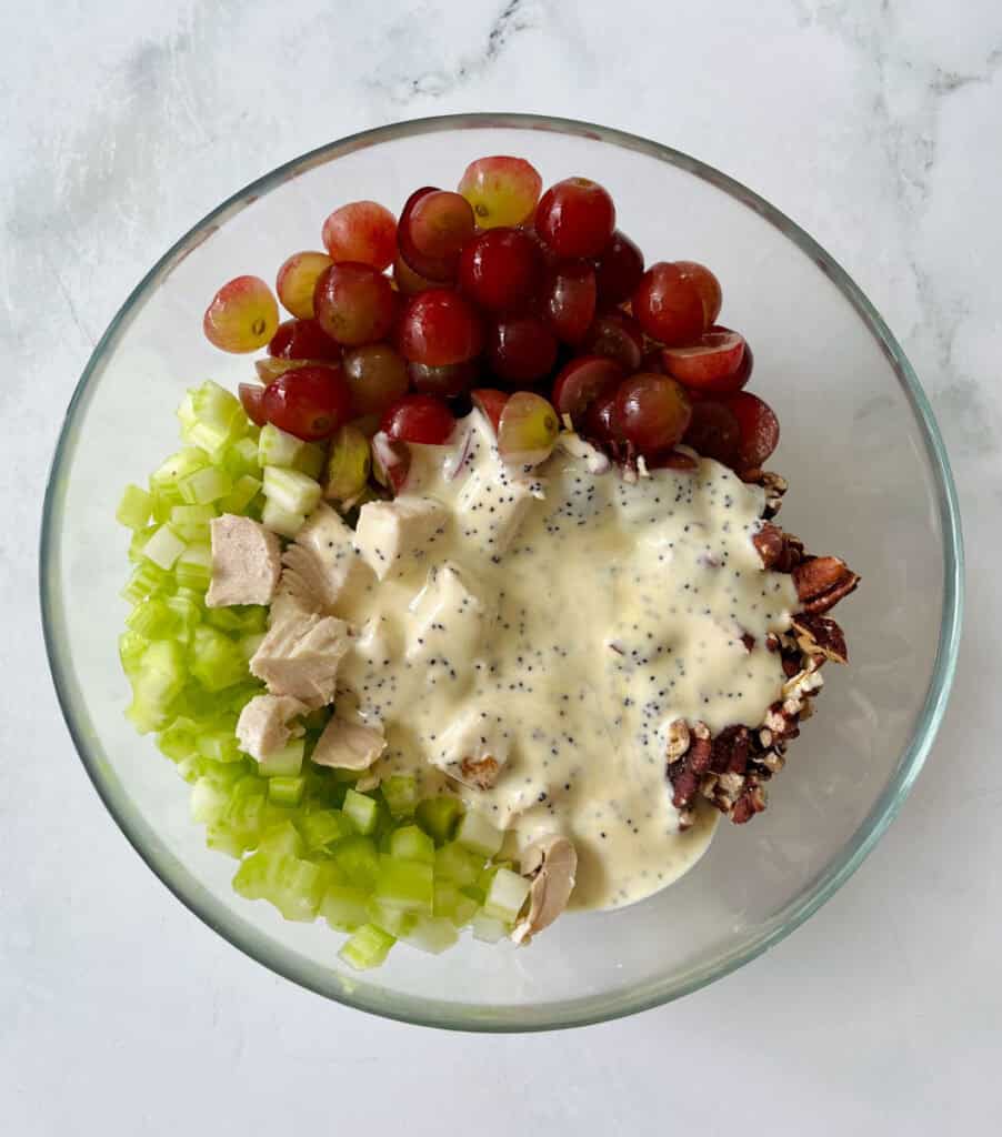A glass bowl filled with grapes, chopped celery, shredded chicken, and a creamy dressing, on a light surface.