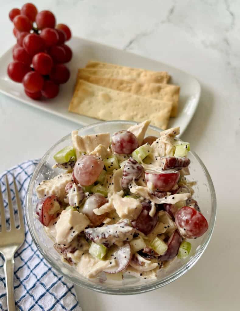 A bowl of chicken salad with grapes, celery, and dressing, accompanied by a plate of crackers and a bunch of red grapes on a white marble surface.