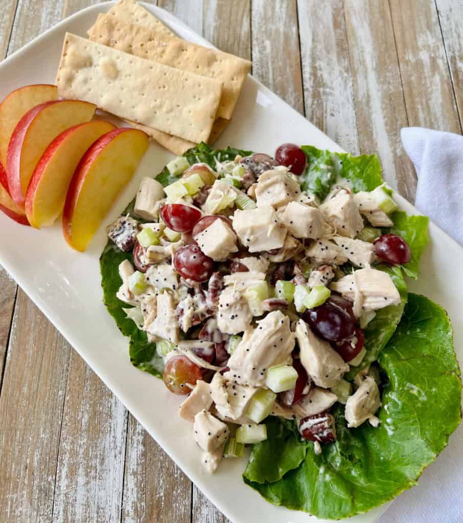 A white plate holds a chicken salad with grapes, celery, and poppy seeds, accompanied by apple slices and saltine crackers, on a wooden table.