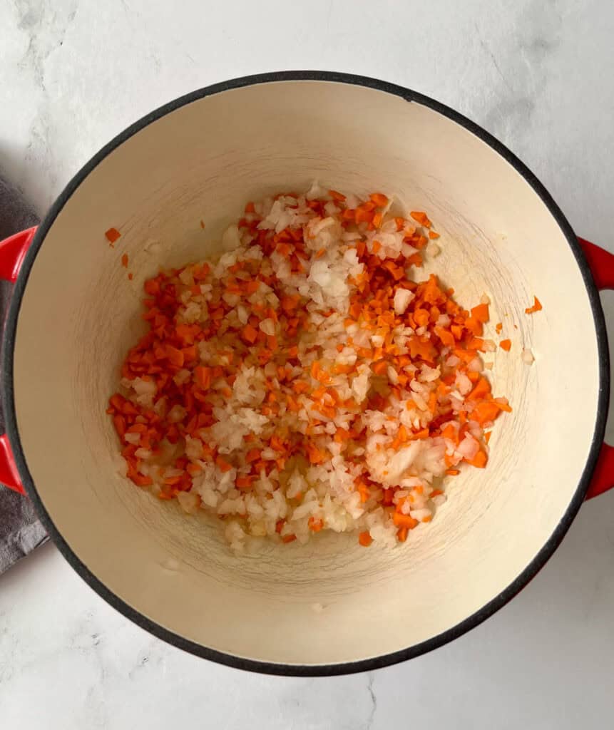 Chopped onions and carrots in a cooking pot on a white marbled surface.