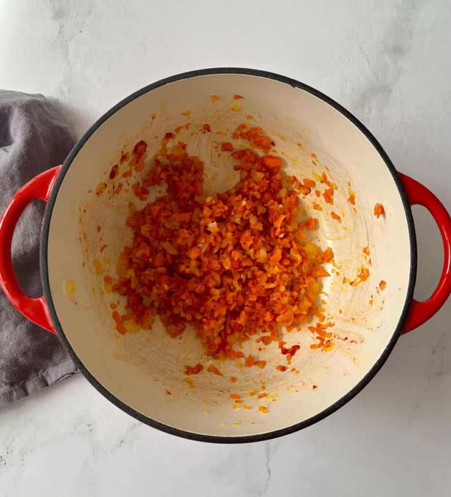 Chopped carrots, onions, garlic, and tomato paste cooking in a red Dutch oven on a white marbled surface.