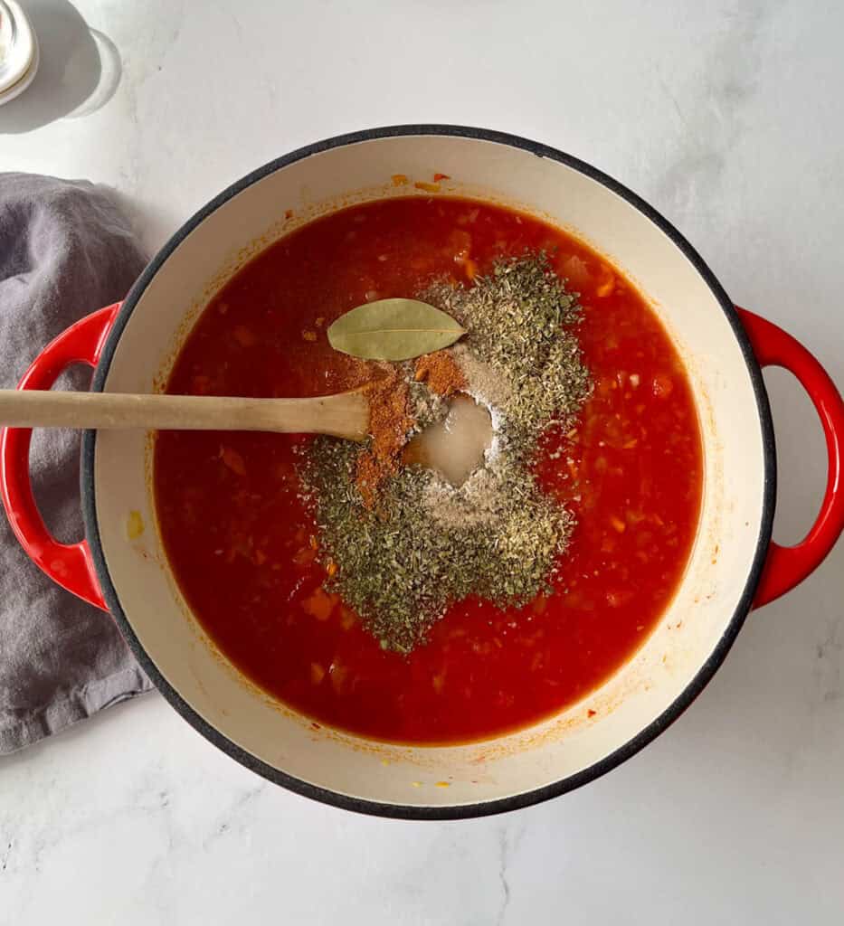 Cooking tomato soup with herbs in a red pot on a white surface.