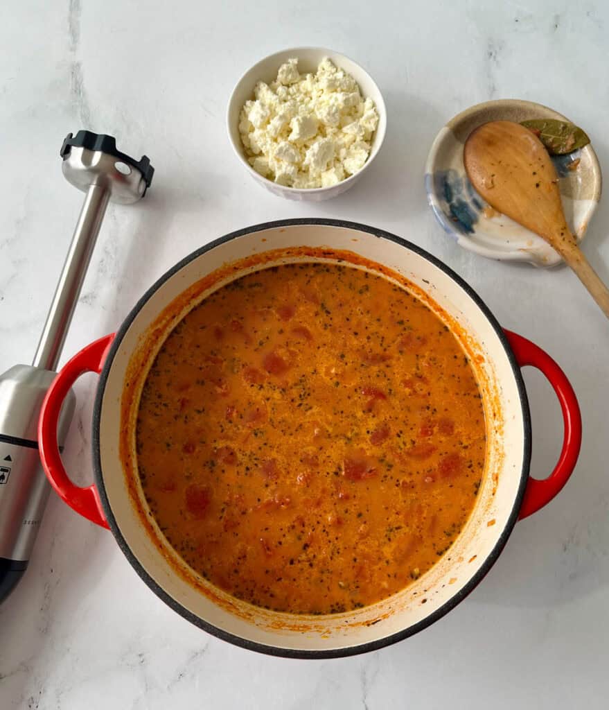 A pot of homemade tomato soup with crumbled cheese and a wooden spoon on a white surface.