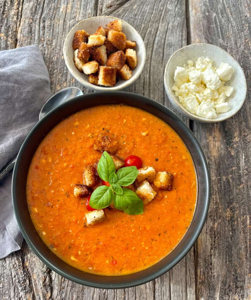 Tomato soup garnished with basil, croutons, and served with cheese and croutons on the side on a wooden surface.