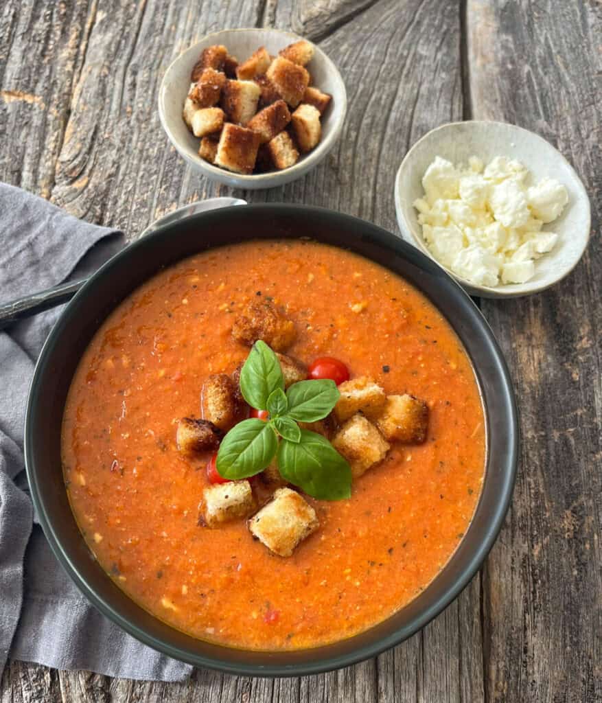 Tomato feta soup garnished with basil and croutons, served in a black bowl on a rustic wooden surface.