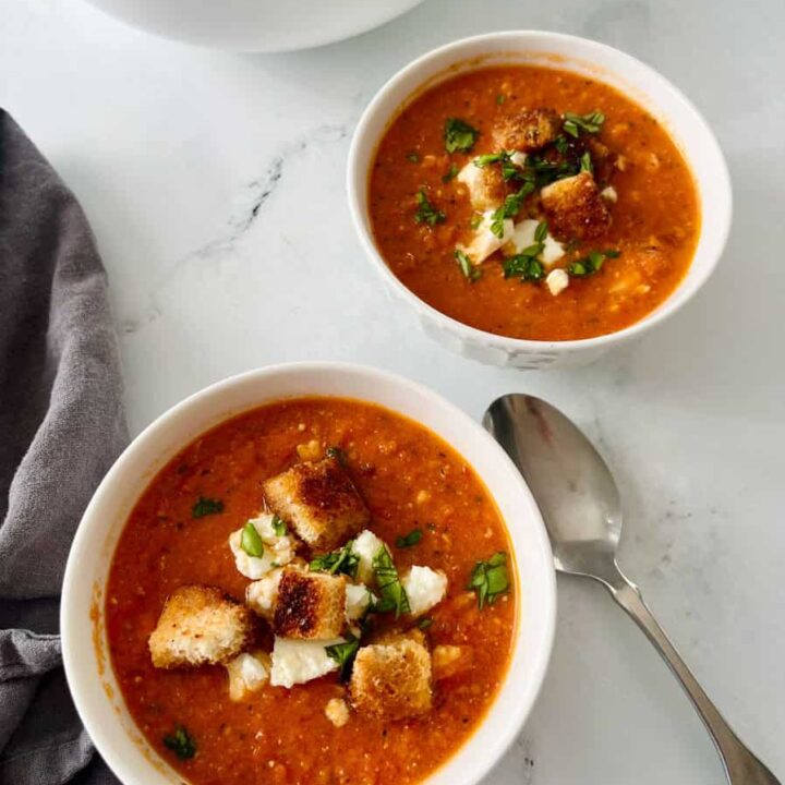Close-up of two bowls of homemade tomato soup garnished with croutons and fresh herbs, served on a white surface with a spoon.