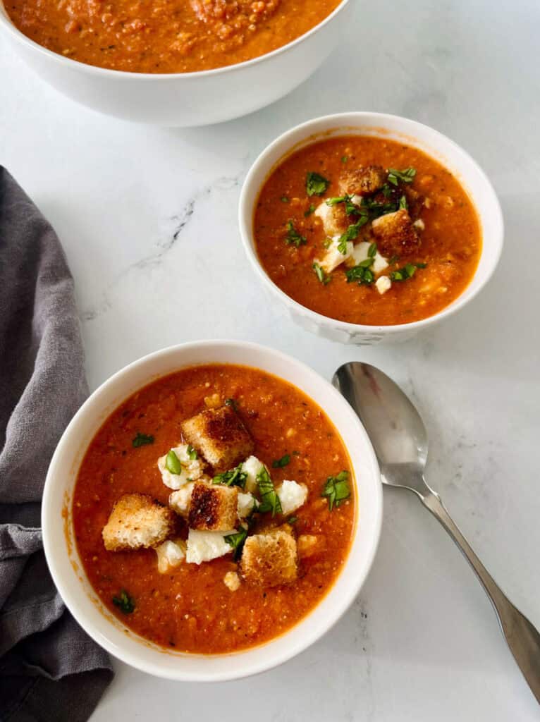 Close-up of two bowls of homemade tomato soup garnished with croutons and fresh herbs, served on a white surface with a spoon.