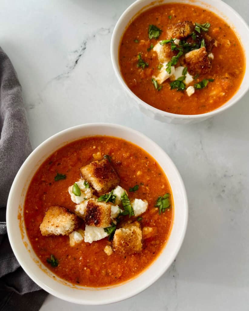 Bowl of tomato soup topped with croutons and fresh herbs on a white marble surface.