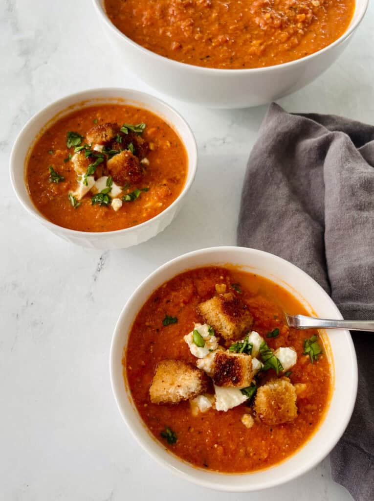 Three bowls of homemade tomato soup garnished with croutons and herbs.