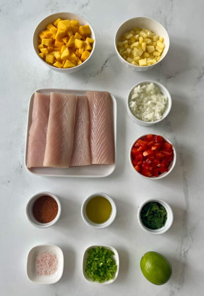 Fresh fish fillets with diced mango, pineapple, onion, peppers, and seasonings for recipe preparation.