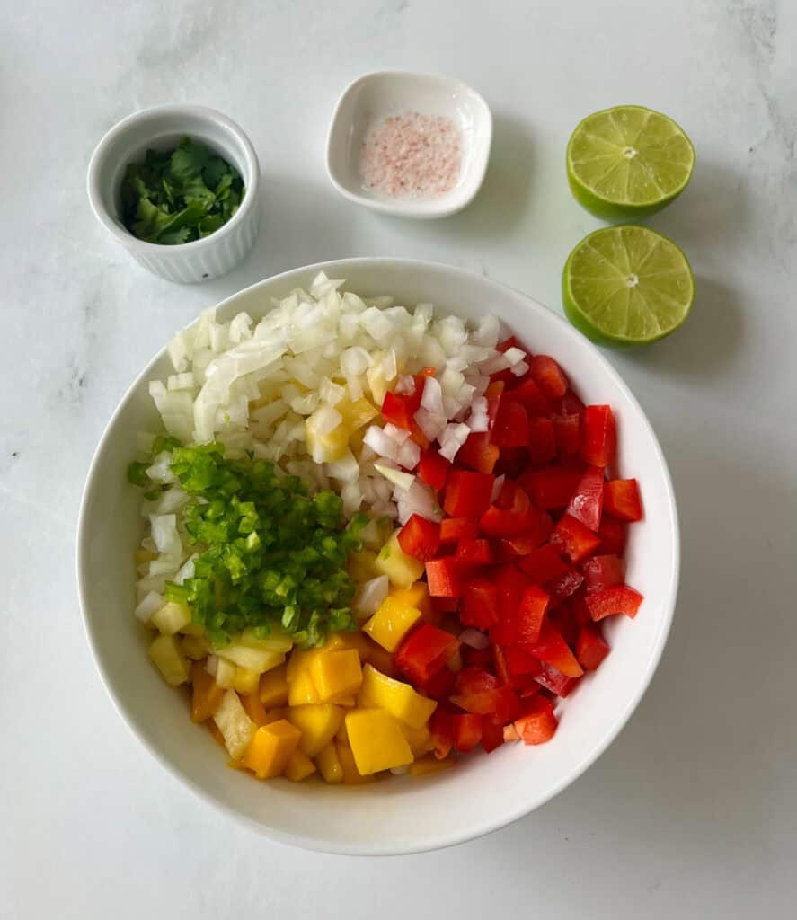 Chopped onions, red bell peppers, pineapple, and mangoes in a bowl with lime and seasonings.