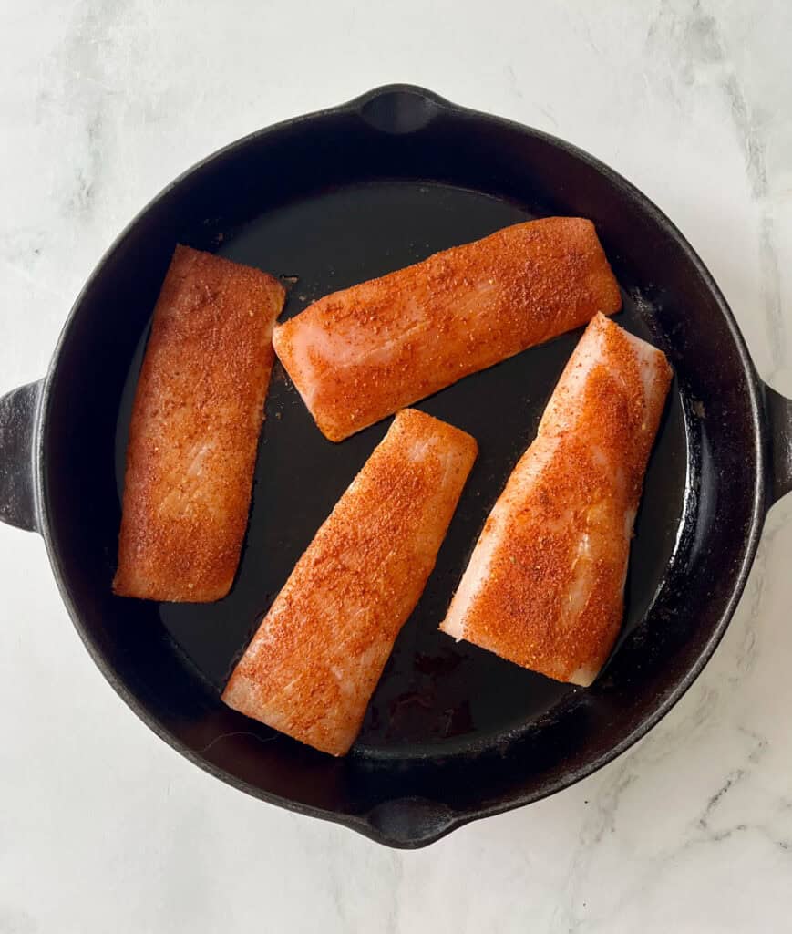Seasoned fish fillets cooking in a black skillet on a white surface.