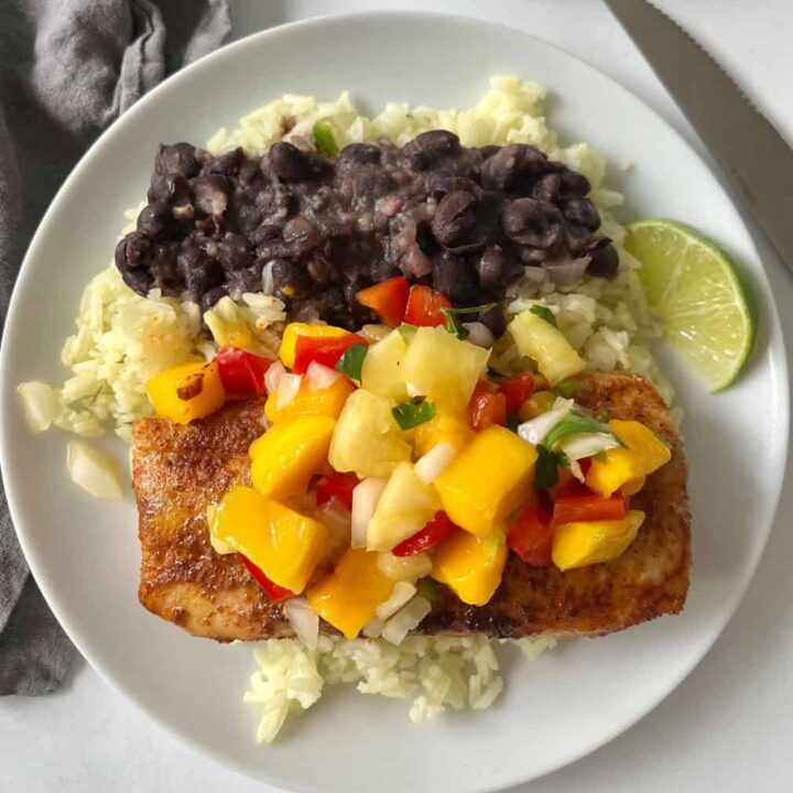 A white plate featuring a fish fillet topped with vibrant mango salsa, black beans, rice, and a lime wedge.