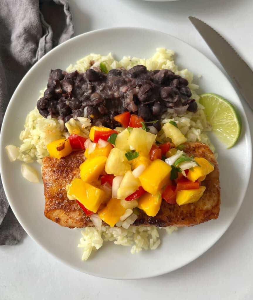 A white plate featuring a fish fillet topped with vibrant mango salsa, black beans, rice, and a lime wedge.