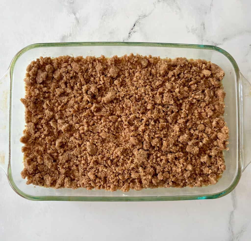 An unbaked coffee cake with a crumble topping in a clear glass dish on a white surface ready to be baked.