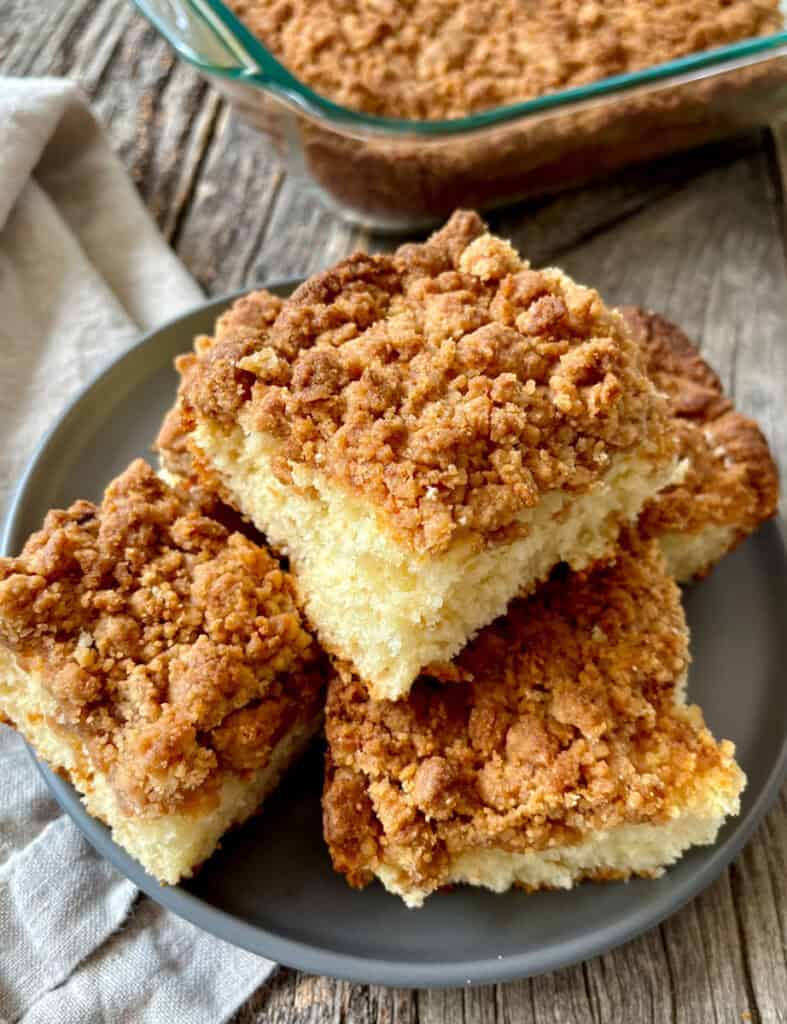 Close-up of freshly baked crumb cake squares with a crumb topping on a dark plate, with a baking dish in the background.