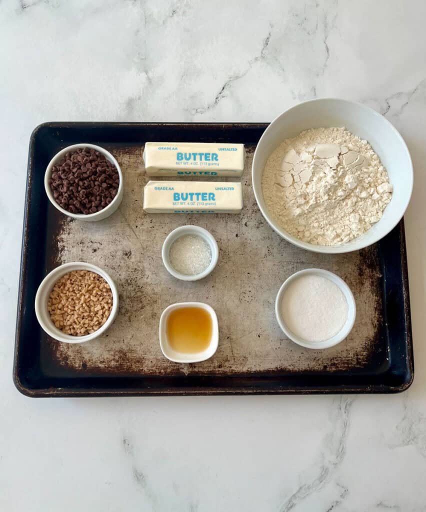 Assorted baking ingredients on a tray including flour, butter, mini chocolate chips, toffee bits, and sugar.