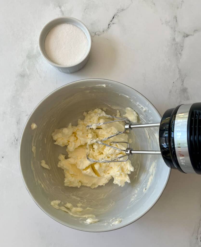 Mixing butter with an electric mixer in a bowl. A small bowl of sugar is in the background.