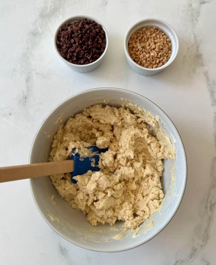 Shortbread cookie dough in a bowl with chocolate chips and toffee bits on a white surface.