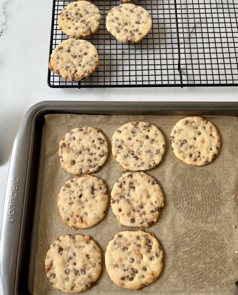 Freshly baked shortbread cookies with chocolate chips and toffee bits cooling on a wire rack and baking sheet.