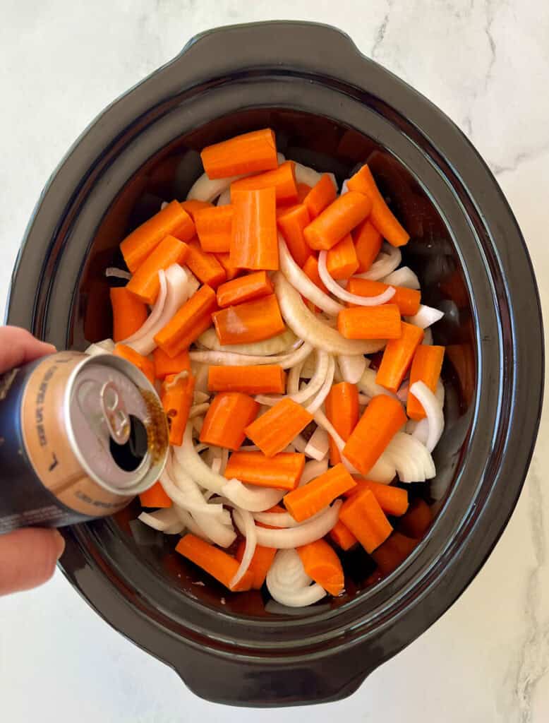 Chopped carrots and onions in a slow cooker with a beer being poured for pot roast.