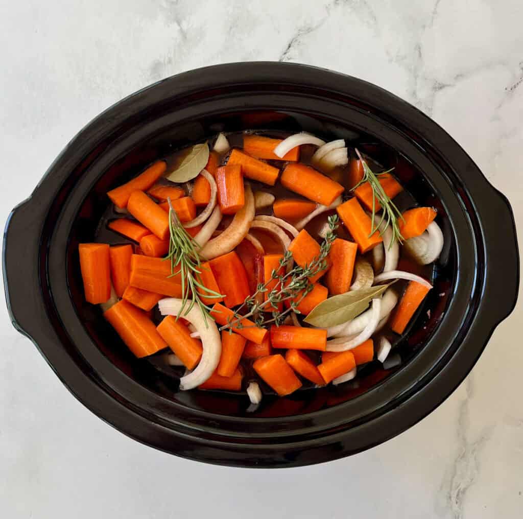 Carrots, onions, garlic, and herbs in a slow cooker ready for cooking.