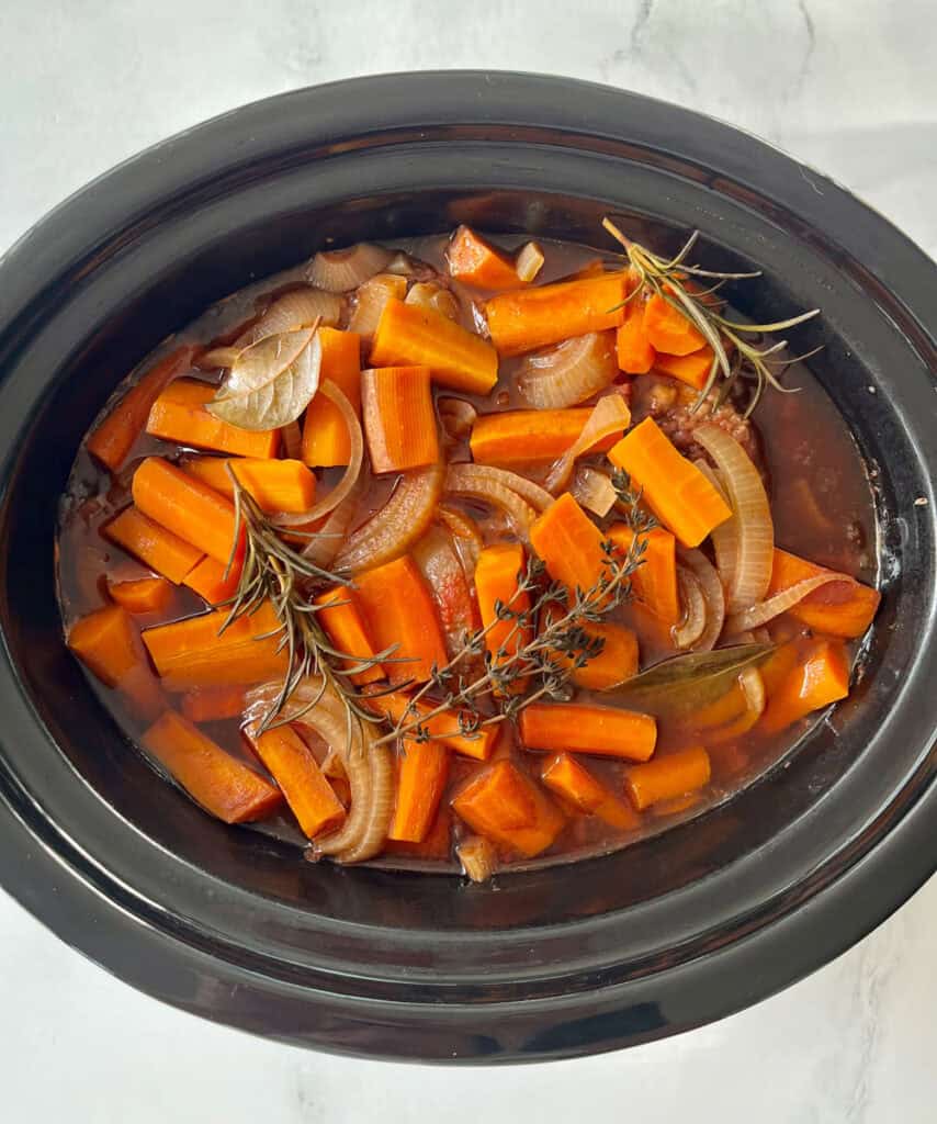 Slow cooker with carrots, onions, beef, and herbs on a marbled surface.