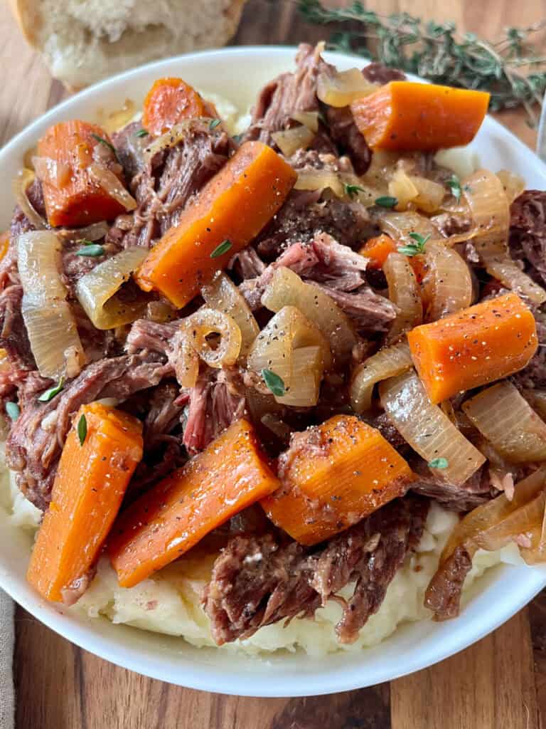 Close-up of beef pot roast with carrots, onions, and herbs served over mashed potatoes.
