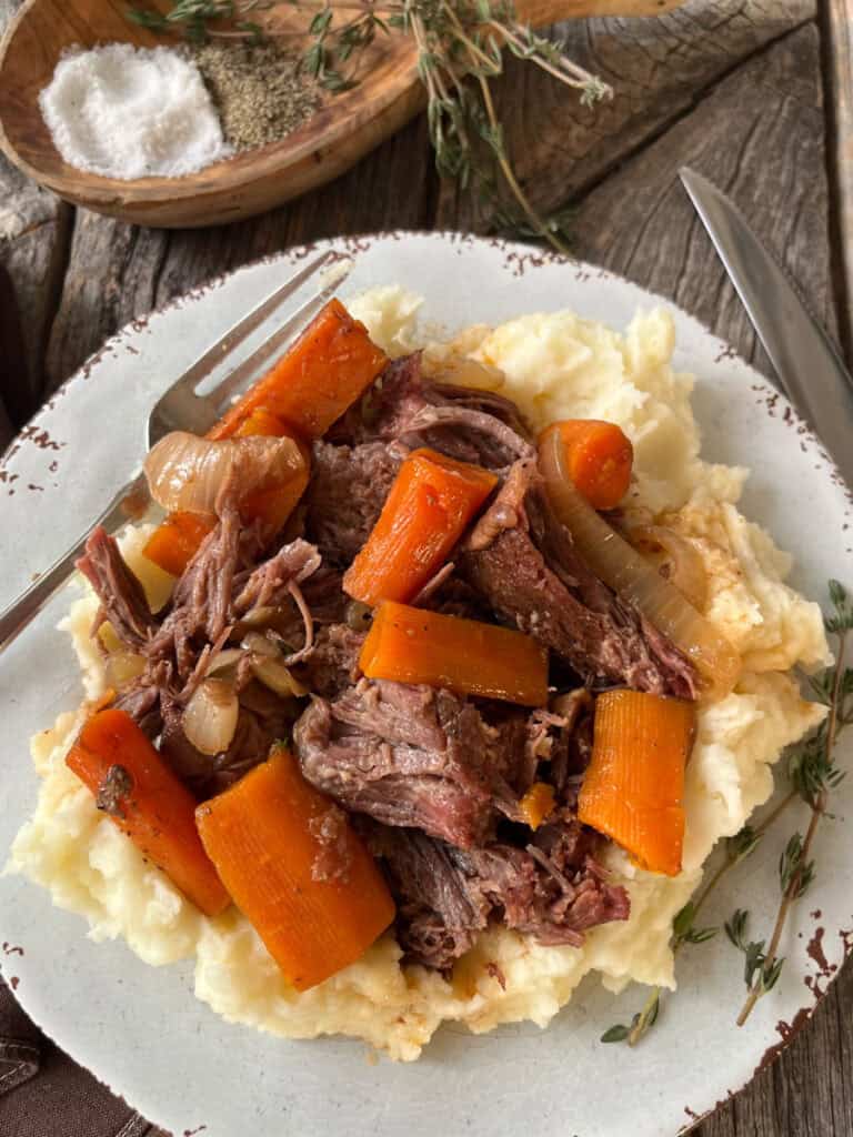 A plate of pot roast with carrots, onions, and mashed potatoes on a rustic wooden table.