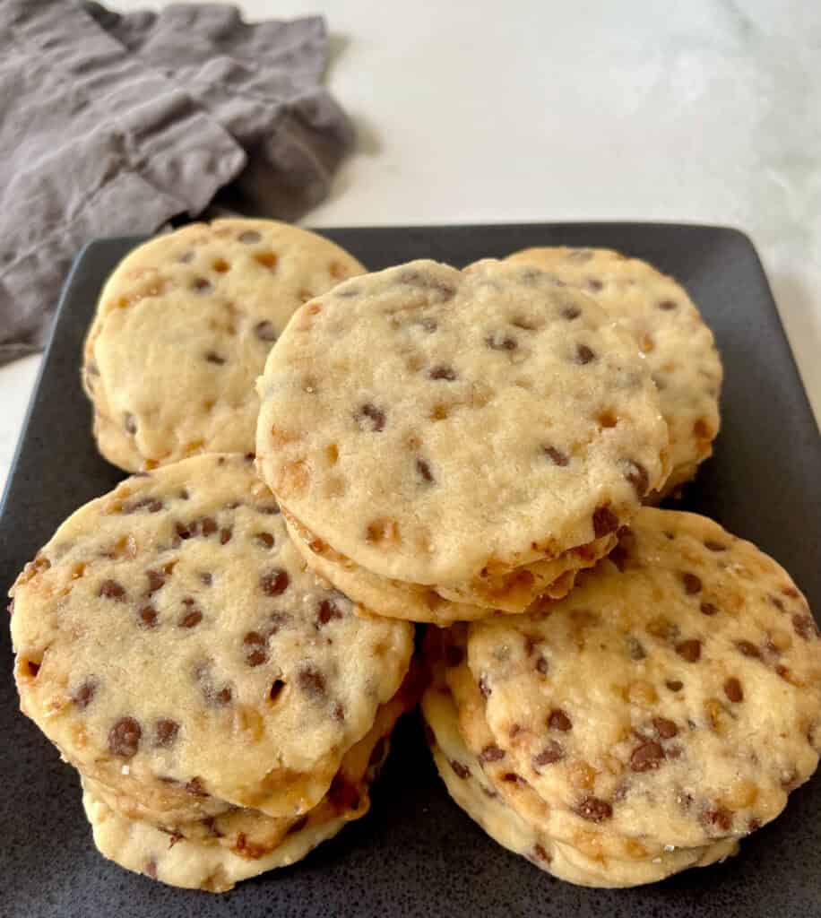 Freshly baked shortbread cookies with chocolate chips and toffee bits stacked on a black plate.