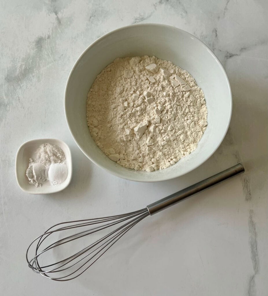 White bowl with flour and a small dish with salt, baking powder, and baking soda and a whisk ready for cooking.