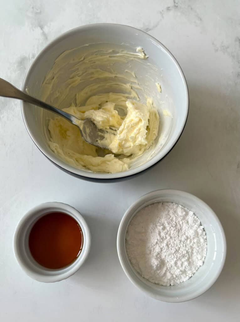 Bowl of creamy butter mixture with a bowl of powdered sugar and a bowl of maple syrup on a white surface.