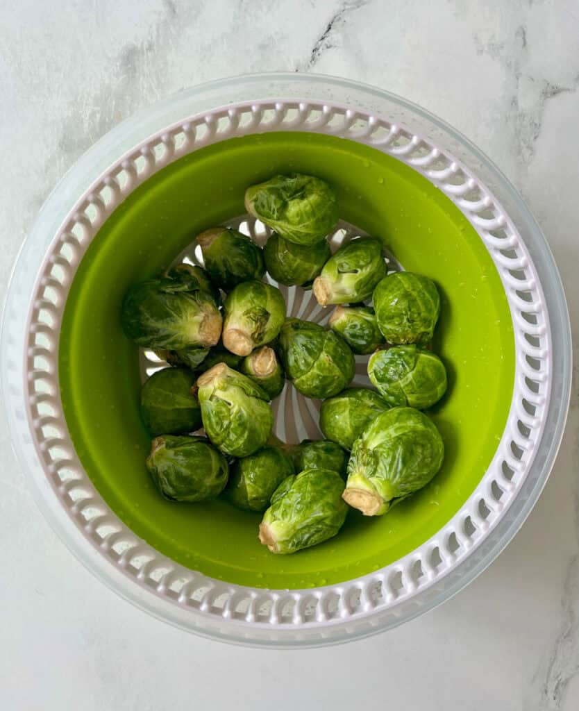 Fresh Brussels sprouts in a green salad spinner on a marble surface.