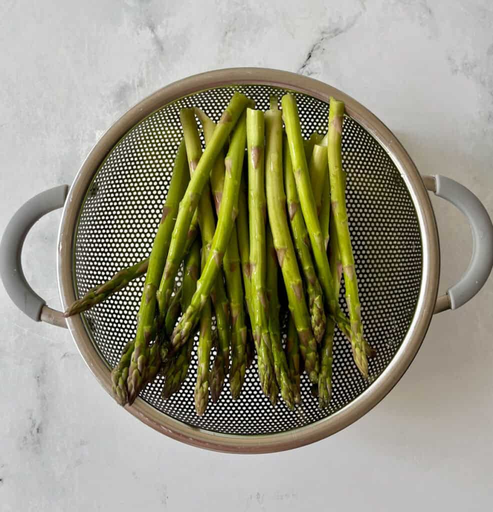 Fresh green asparagus spears in a metal colander on a light surface.