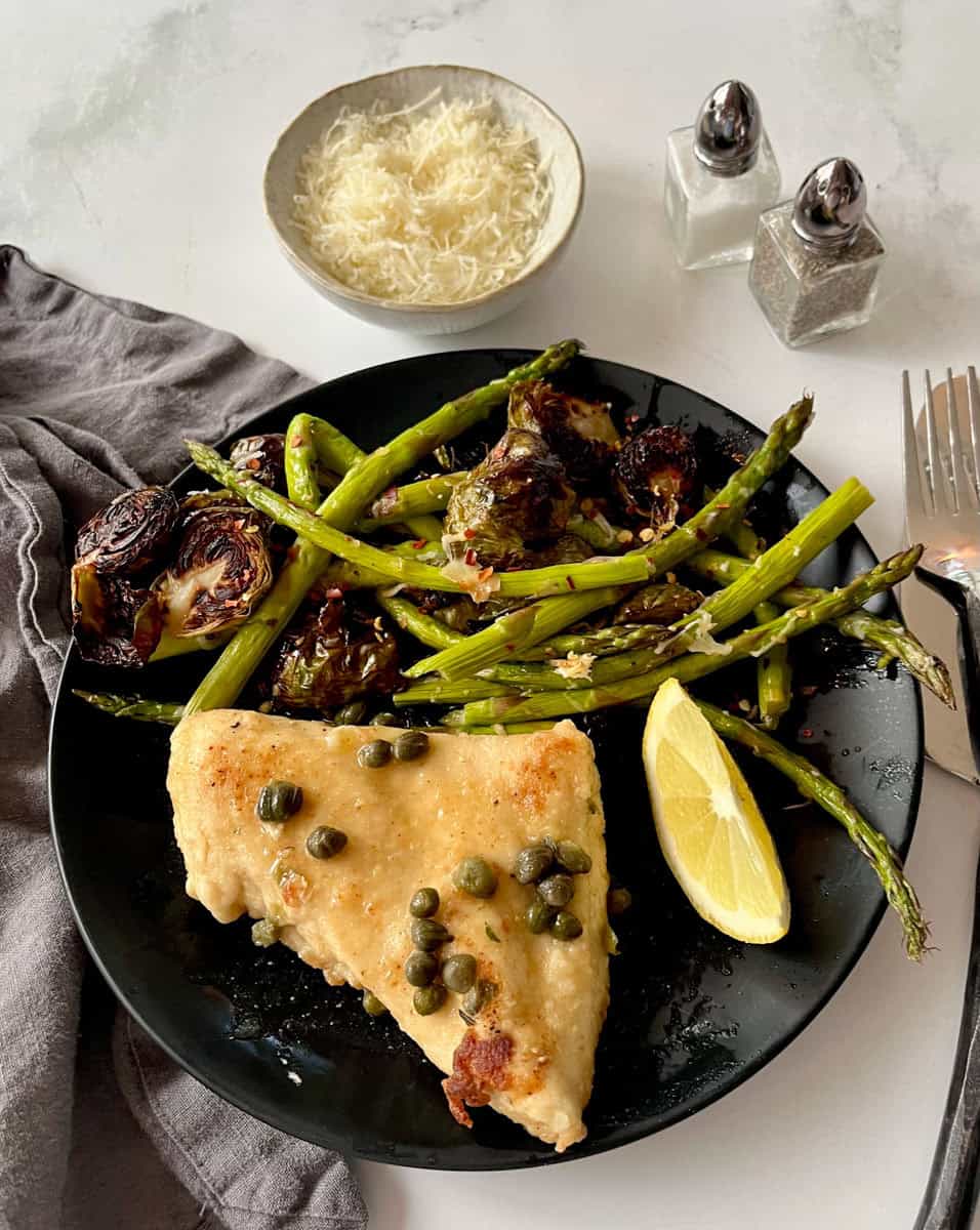 Chicken with capers accompanied by Brussels sprouts and green asparagus, lemon wedge, grated cheese, and seasonings on a black plate.