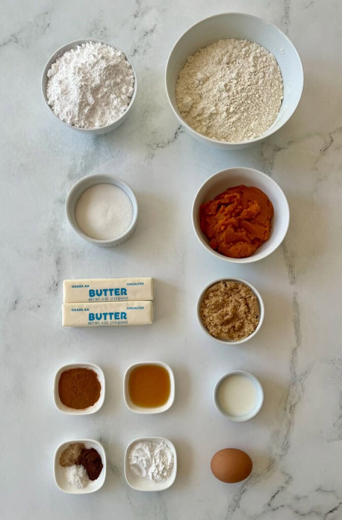 Assorted baking ingredients including flour, sugar, butter, pumpkin, and spices on marble surface.