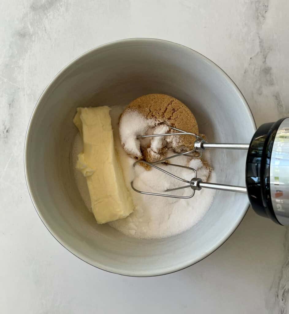 A mixing bowl containing sugar, butter, and brown sugar, with a hand mixer attachment.